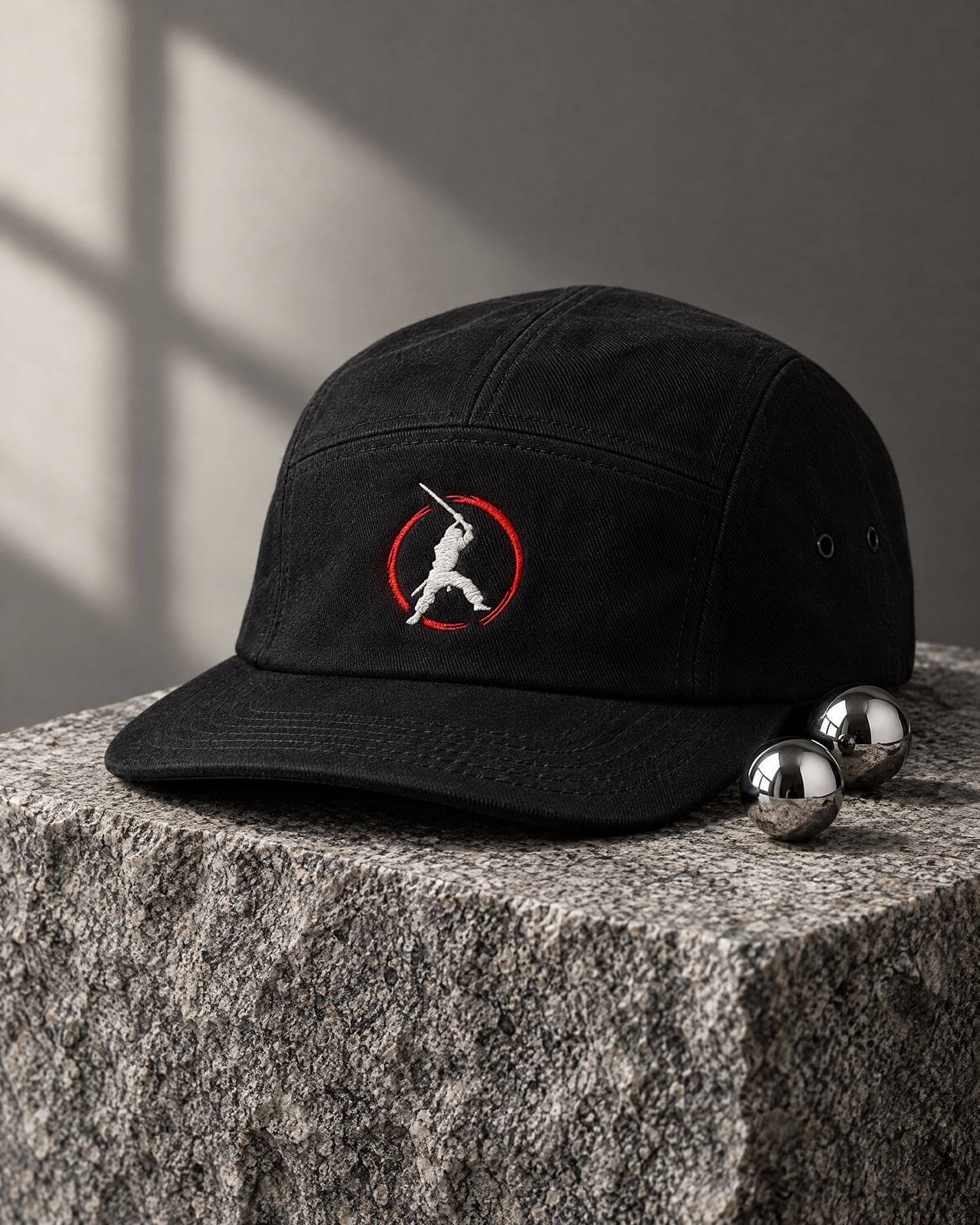 Concept mockup of a black five-panel cap with an embroidered ninja in a red ring, on granite beside two steel pinballs