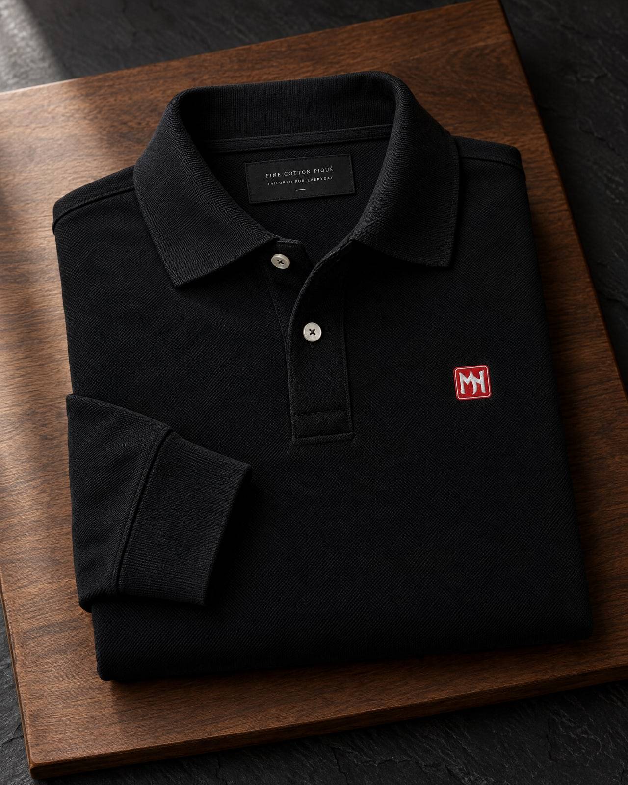Concept mockup of a folded black long-sleeve collared shirt with a small red MN seal embroidered on the left chest, on walnut and slate