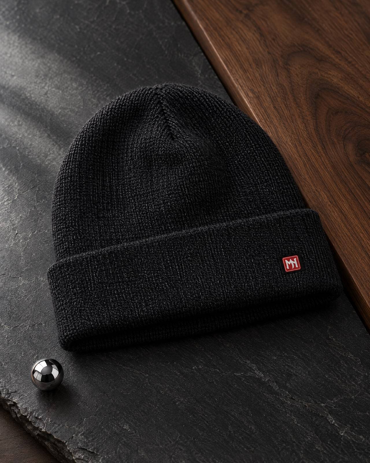 Concept mockup of a folded charcoal fine-knit beanie with a tiny red MN seal label on the cuff, on slate beside a steel pinball