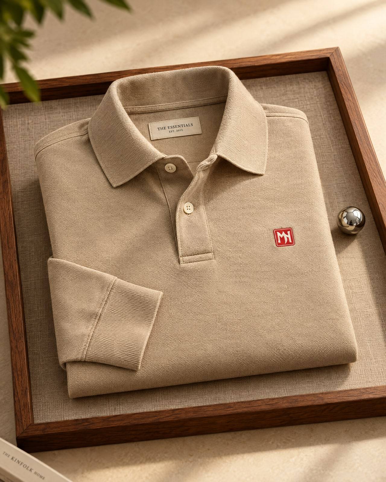 Concept mockup of a folded sand khaki long-sleeve polo with a small red MN seal on the left chest, on a linen tray